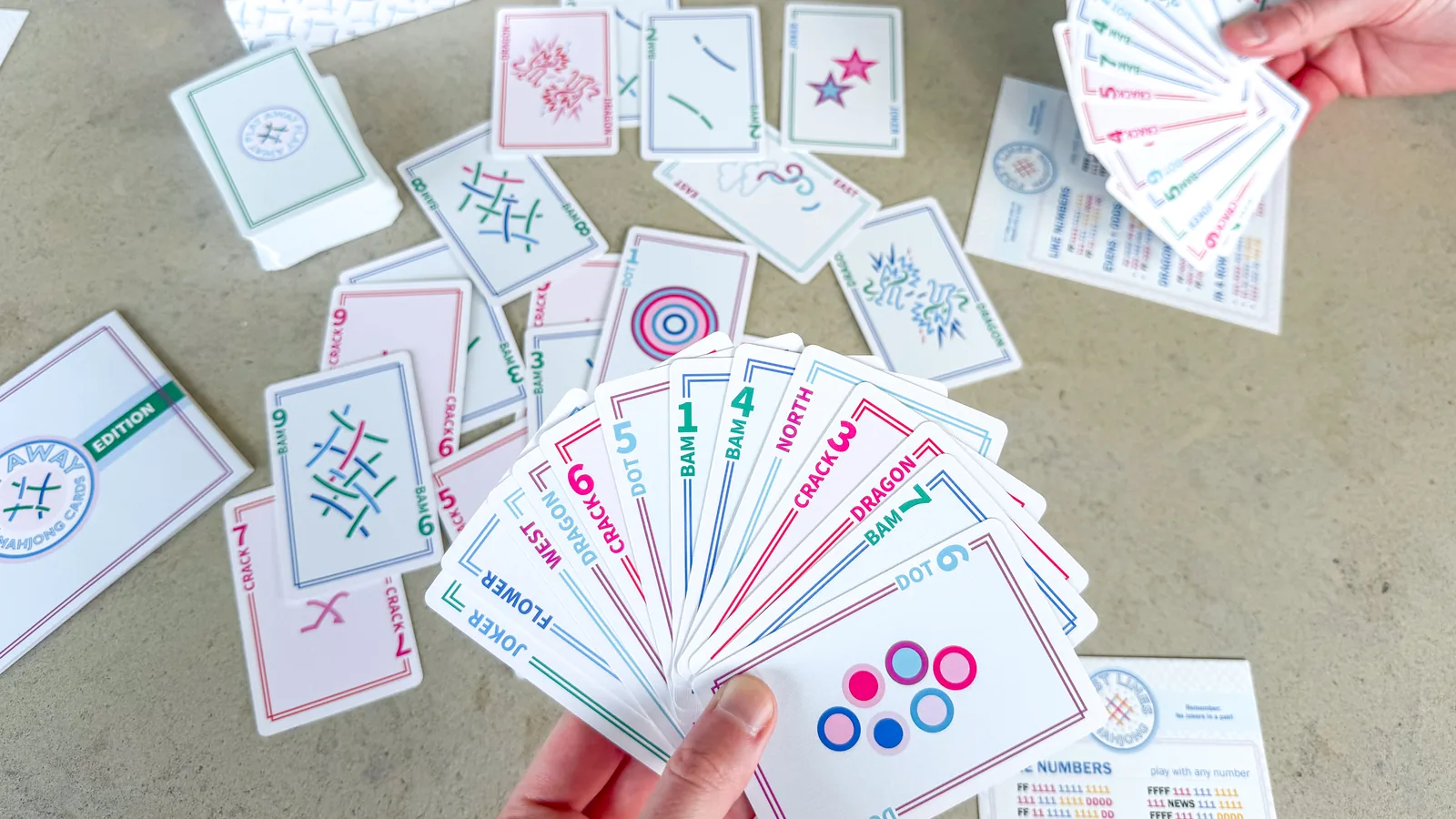 A hand fanning a hand of Play Away cards over a scatter of cards and a First Lines card on a concrete table