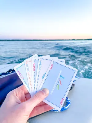 A hand holding a fan of five Play Away cards on a boat, with open water and a low sun behind