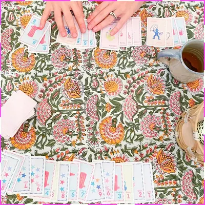 Overhead of hands dealing Texas Edition cards on a floral block-print tablecloth beside a mug of tea