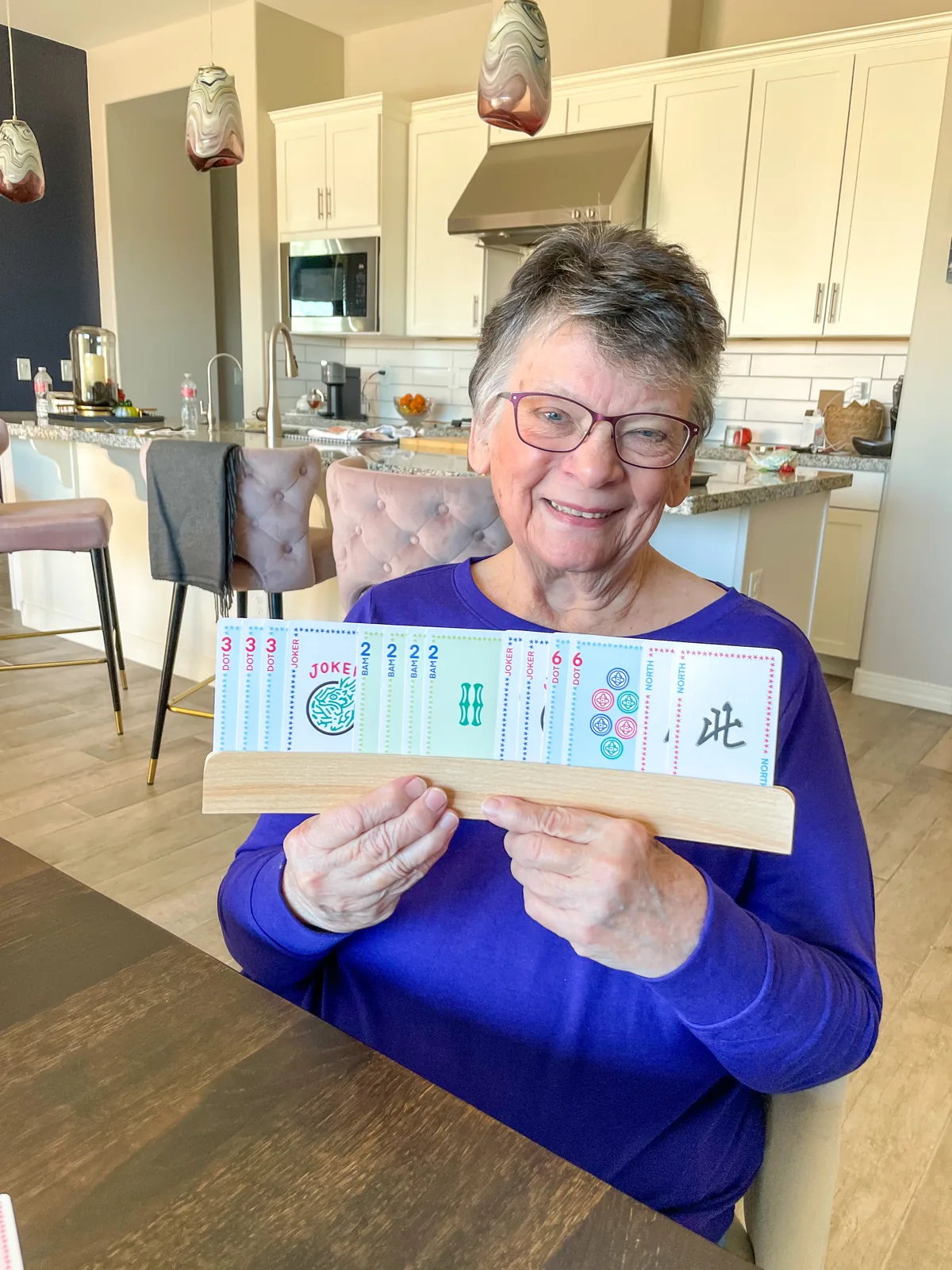 A smiling woman at a kitchen table holding up a wooden rack of Classic Edition cards