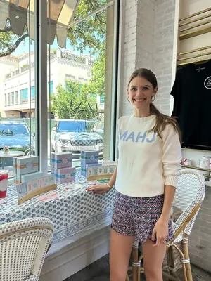 A woman in the White/Blue MAHJ Knit Sweater standing beside a table of Play Away card boxes in a shop window